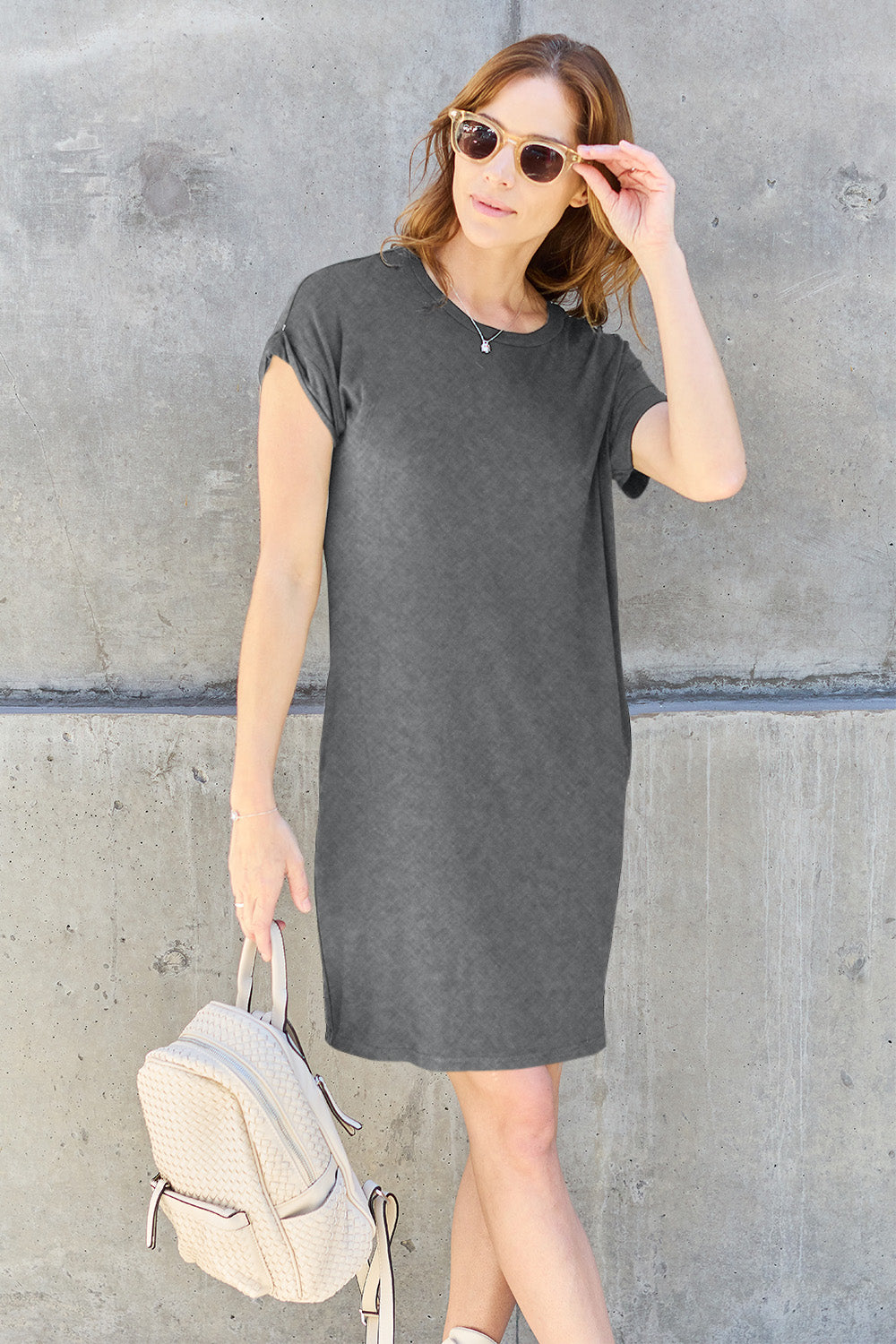 A woman wearing a dark olive-green Basic Bae Full Size Round Neck Short Sleeve Dress with Pockets and sunglasses stands against a concrete wall, holding a light-colored backpack.