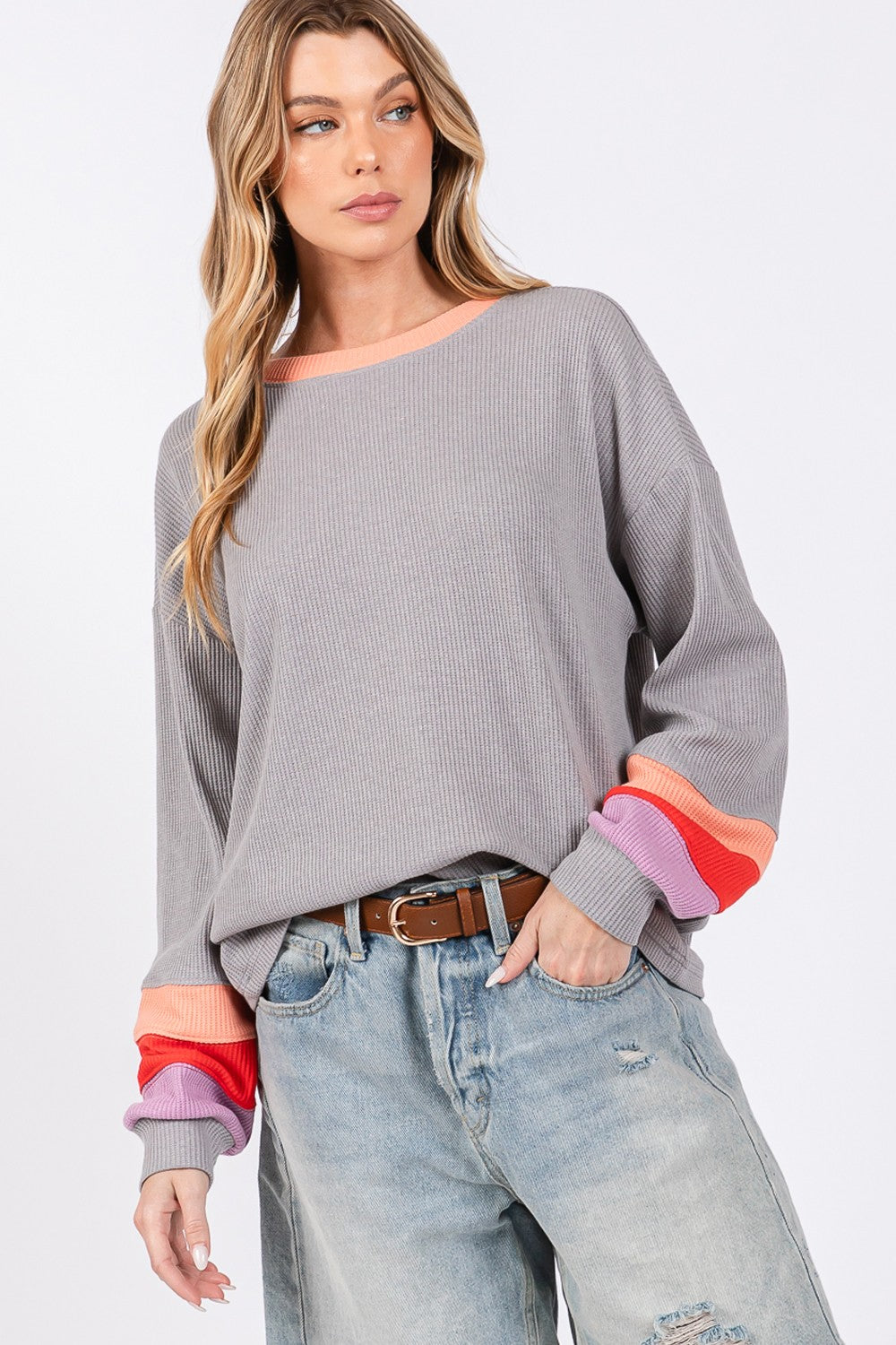 A woman wearing the SAGE + FIG Color Block Waffle Knit T-Shirt stands with one hand on her gray hat. The casual top, adorned with colorful block raglan sleeves, pairs effortlessly with denim shorts for a chic look.