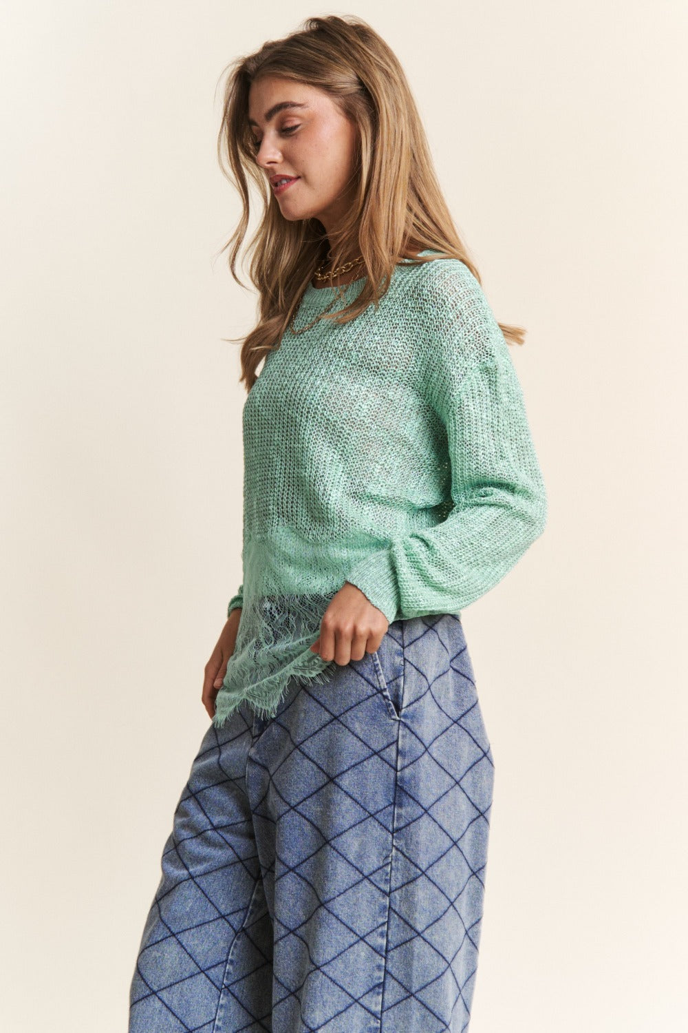 A woman with long hair is wearing the J.NNA Lace Detail Long Sleeve Knit Top in a versatile light green, paired with a lace trim shirt underneath and patterned blue pants. She has a neutral expression and her right hand near her face.