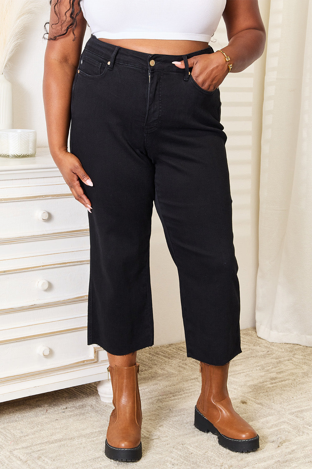 A person wearing Judy Blue Full Size High Waist Wide Raw Hem Cropped Jeans in black and a white lace top stands next to a white dresser adorned with a vase and candle.