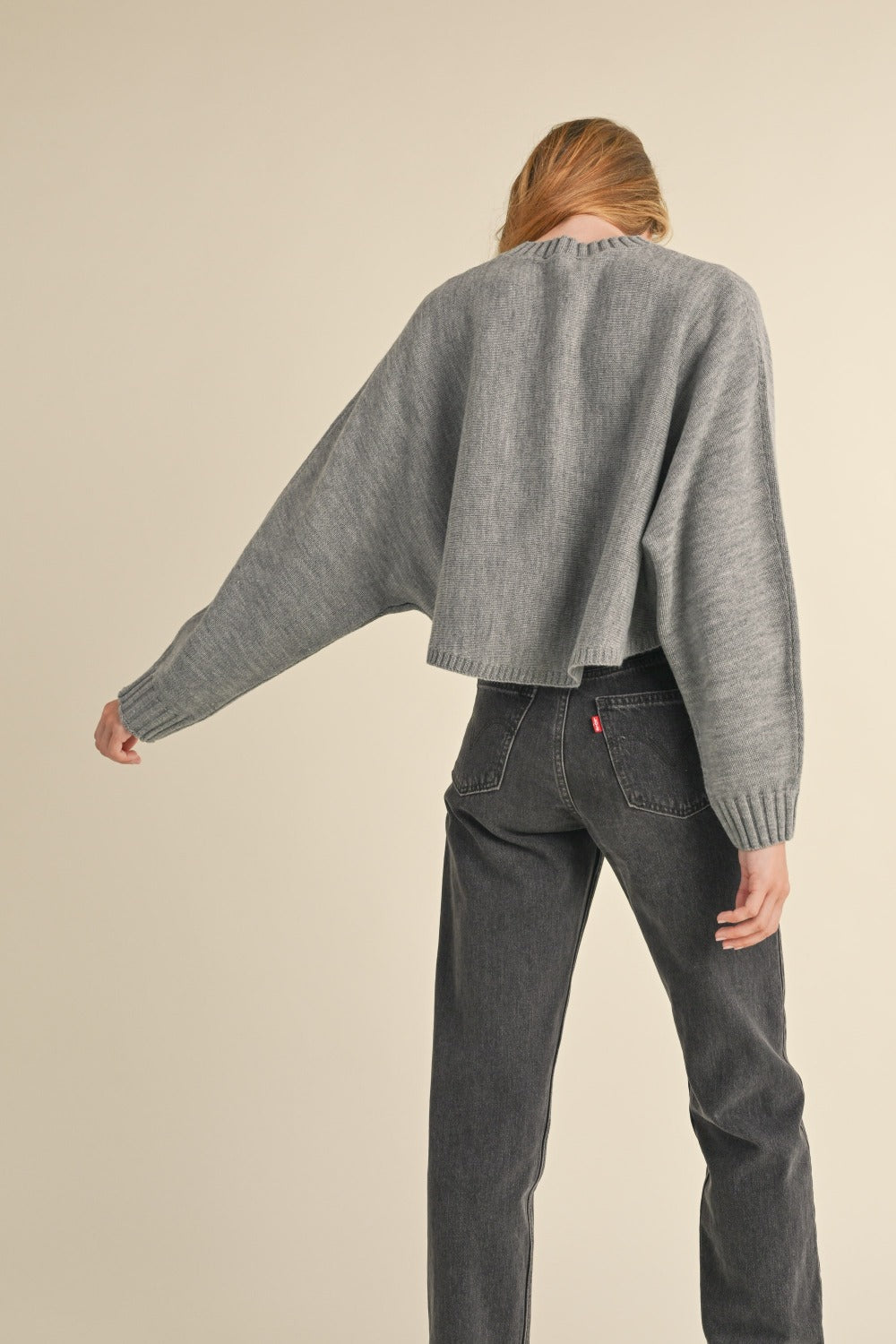 A person with long, light brown hair is facing away, wearing a Mable Round Neck Dolman Sleeve Cropped Sweater in grey paired with dark jeans, against a neutral background.