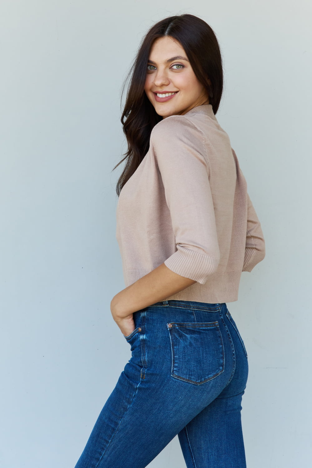 Woman with long dark hair wearing the versatile Doublju My Favorite Full Size 3/4 Sleeve Cropped Cardigan in Khaki over a white top and blue jeans, standing against a plain background.