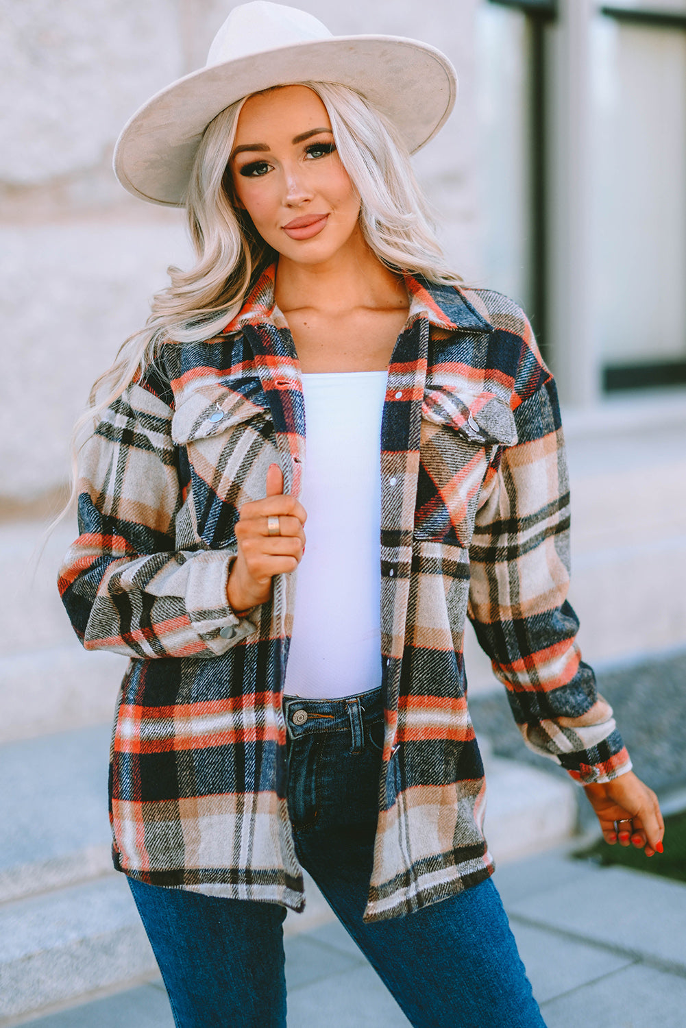 A person wearing a Brown Plaid Button Up Long Sleeve Flannel Shacket layered over a white top with ripped jeans and holding a brown handbag stands against a textured gray wall.