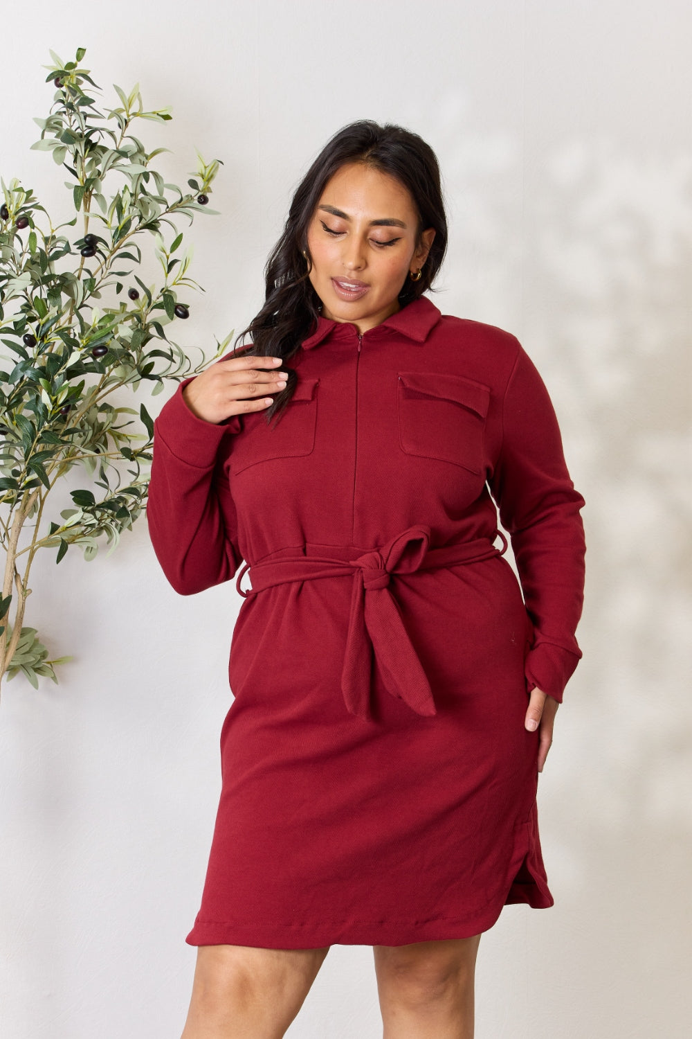 A woman wearing the Culture Code Full Size Tie Front Half Zip Long Sleeve Shirt Dress stands in front of a plain background next to a potted plant.