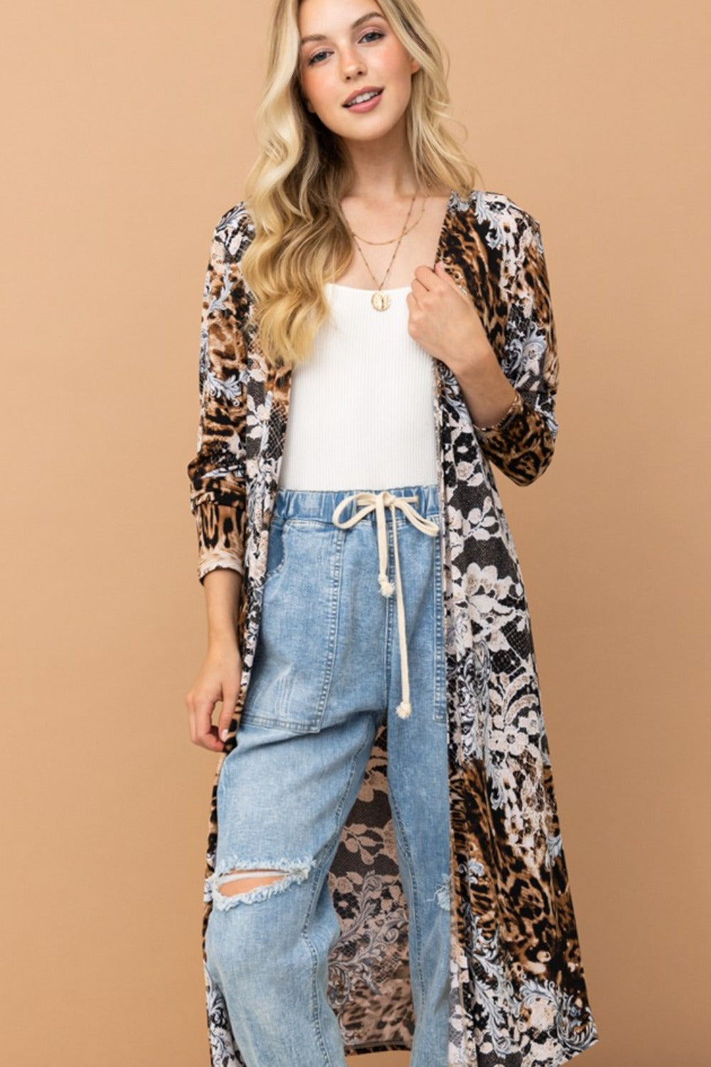 A woman with blonde hair wears a white tank top, the And The Why Leopard Kimono Open Front Longline Cardigan, light blue drawstring jeans, and sandals, standing against a beige background.