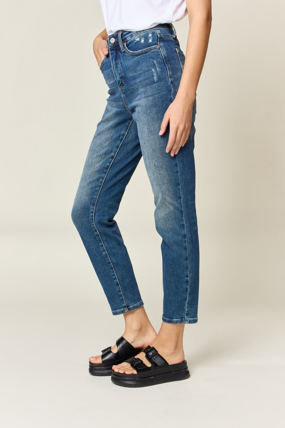 Person wearing Judy Blue Full Size Tummy Control High Waist Slim Jeans in blue and a white shirt, with their hands in their pockets. They are standing indoors on a polished concrete floor with a wooden cabinet and plant in the background.