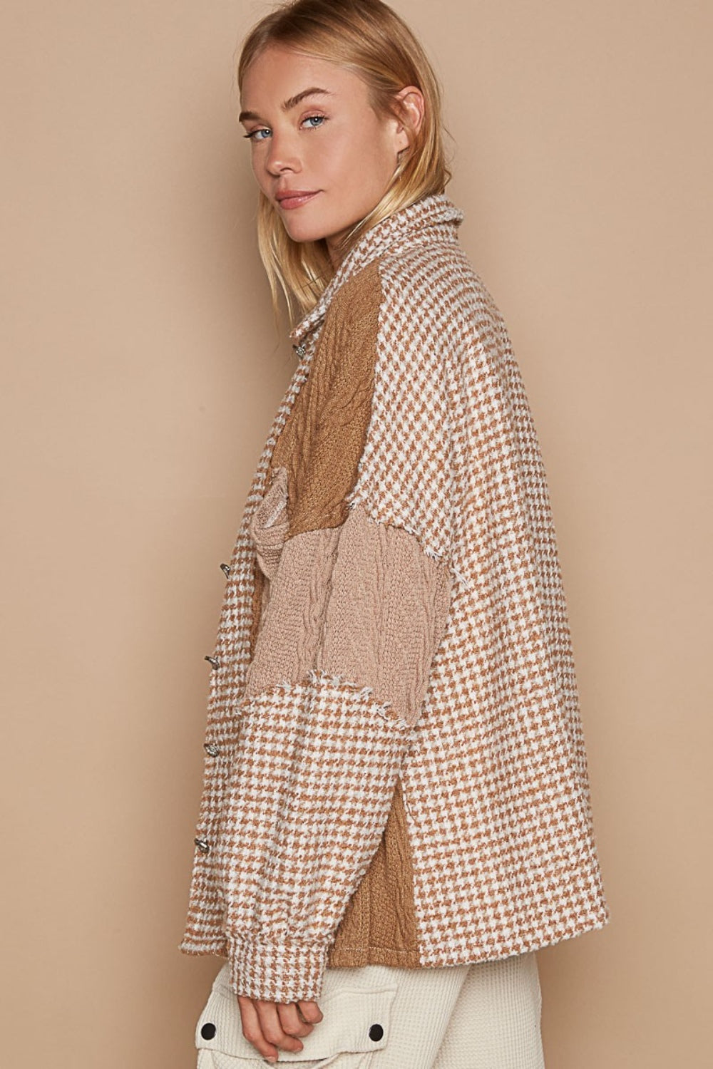 A woman with long blonde hair wears the POL Houndstooth Contrast Cable Knit Pocket Shacket, featuring a patchwork design in various shades of brown and beige, over a white V-neck shirt and paired with light-colored pants. She is standing against a neutral background.