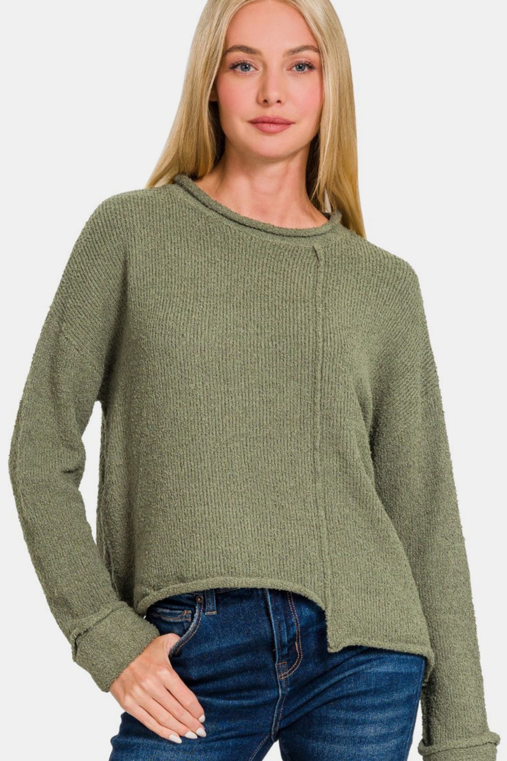 A woman with long blonde hair, dressed in the trendy Zenana Asymmetric Hem Drop Shoulder Sweater in green and blue jeans, poses against a plain background.