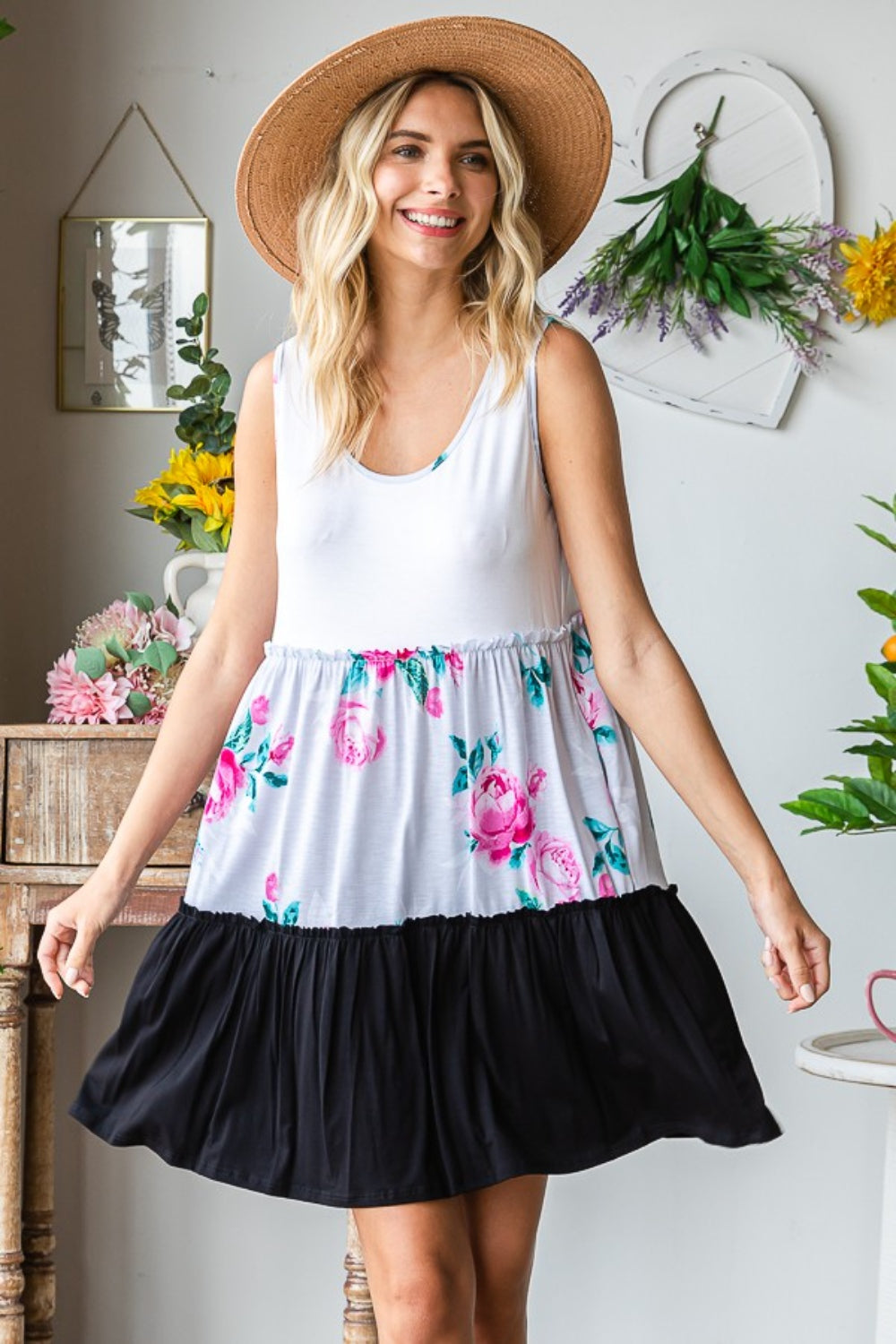 The woman wears the Heimish Full Size Scoop Neck Frill Tiered Dress, showcasing its versatile design with floral and black color blocks. She is indoors, beside a wooden table adorned with plants and wall art.