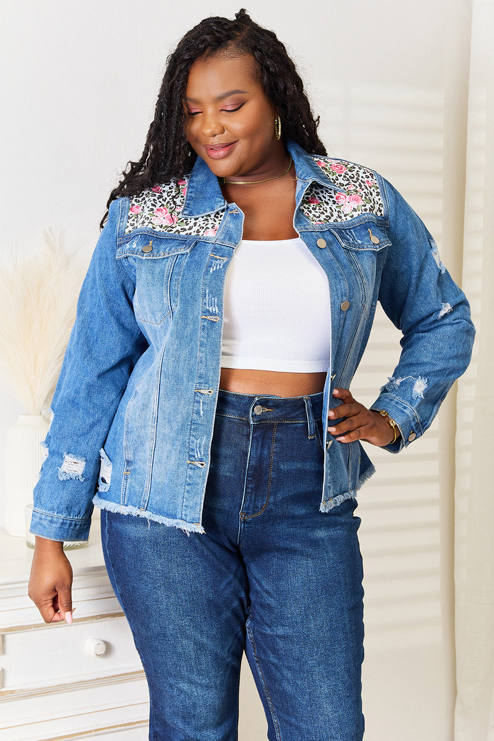 A person smiles while wearing the Baeful Leopard Floral Distressed Raw Hem Denim Jacket with embroidered patches, paired with a white shirt and blue jeans, as they stand indoors near a white piece of furniture.