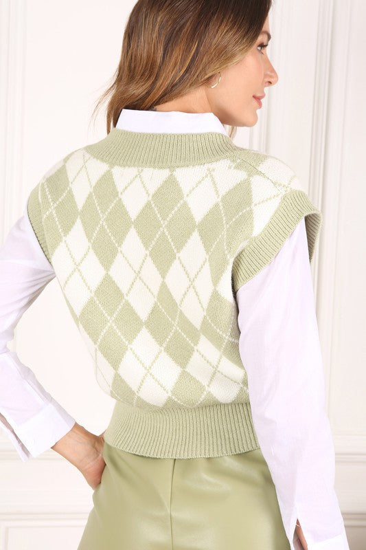 A person wearing a knitted argyle sweater vest in light green over a white shirt stands against a neutral background.