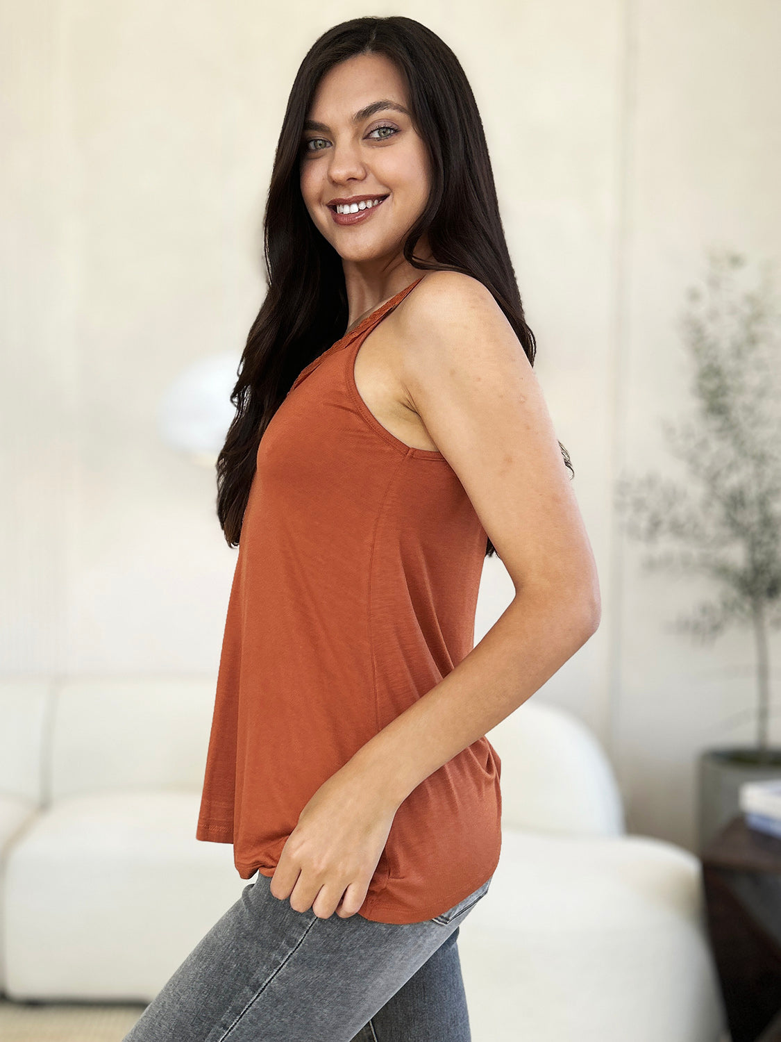 A woman with long hair wearing sunglasses, a slightly stretchy pink "Basic Bae Full Size Lace Detail V-Neck Cutout Cami," and basic style blue jeans stands against a concrete wall, looking to the side with one hand touching her upper chest.