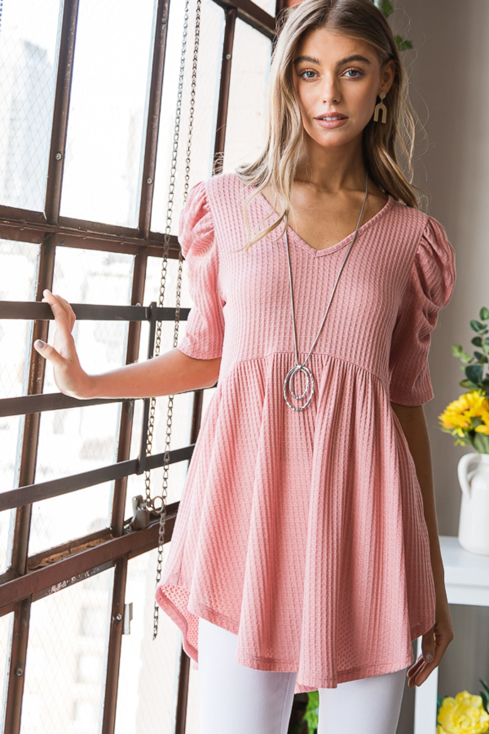 A woman in a Heimish Full Size Waffle Knit V-Neck Babydoll Top with short-sleeves and a long necklace, standing by a window with a neutral expression; the perfect cozy wardrobe addition.