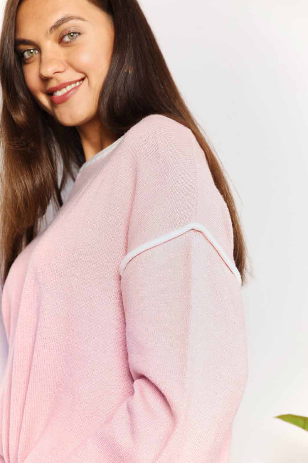 A woman with long curly hair is wearing the Angel Wings Contrast Detail Dropped Shoulder Knit Top in pink, paired with blue jeans, standing against a light background.