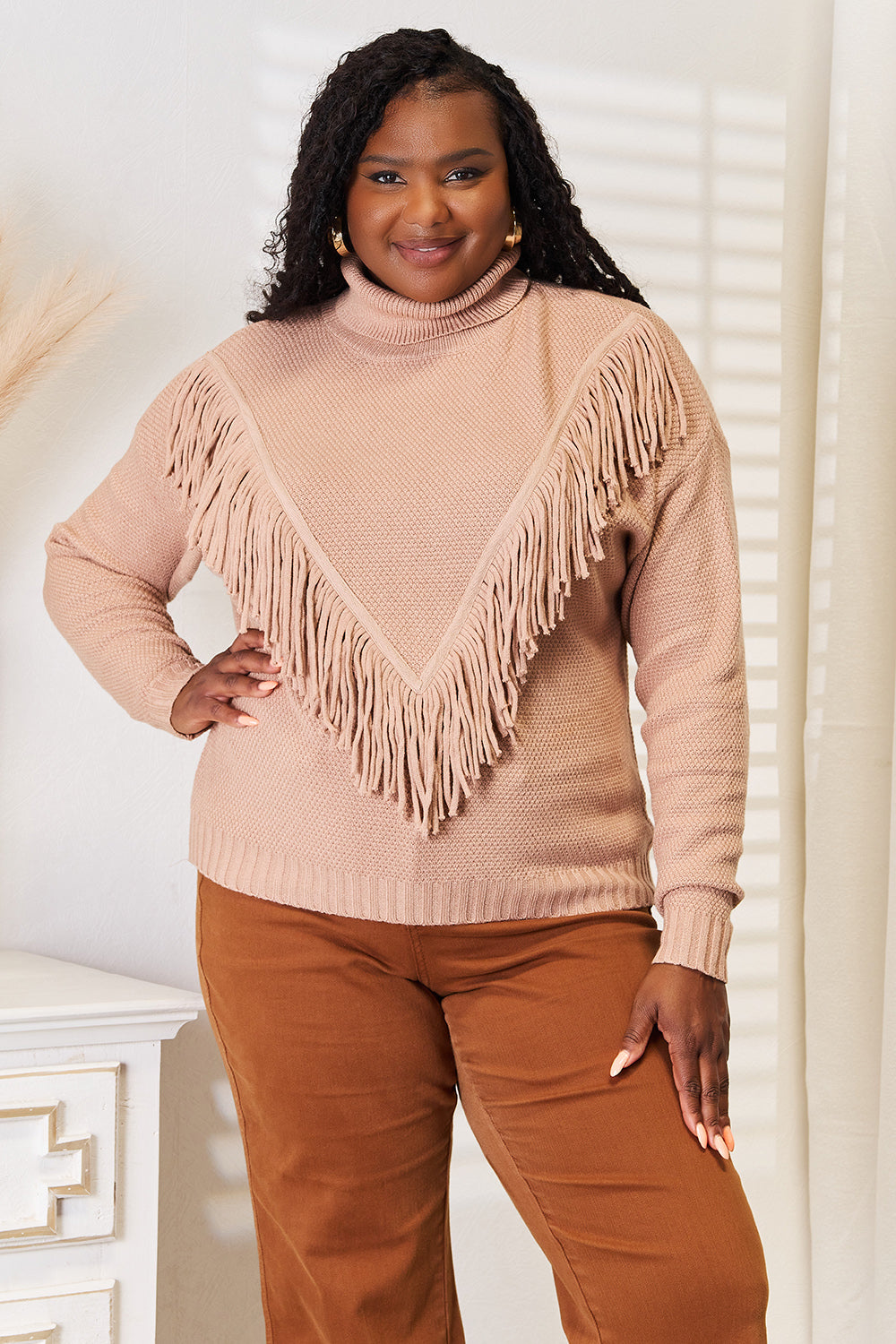 A woman wearing the Woven Right Turtleneck Fringe Front Long Sleeve Sweater, her winter essential, pairs it effortlessly with brown pants. She stands indoors with a neutral expression, embodying cozy sophistication.