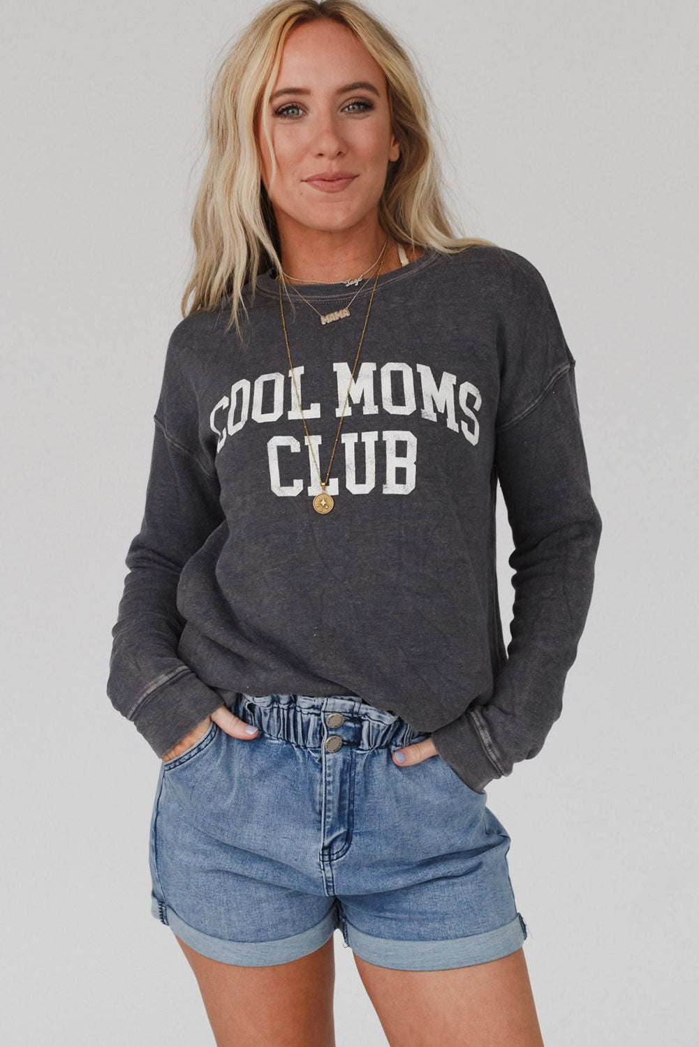 A person with long blond hair wearing a gray COOL MOMS CLUB drop shoulder sweatshirt and relaxed-fit blue jeans is seen from the back, standing near a pumpkin and a window.