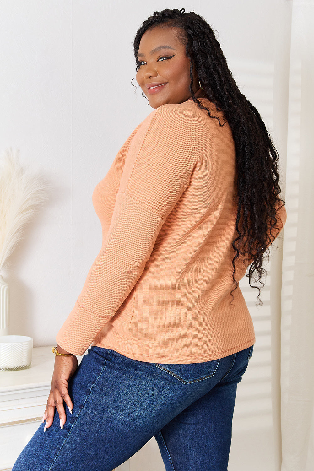 A woman stands indoors, wearing a Basic Bae Half Button Long Sleeve Top in peach and blue jeans, with her hand resting on her hip, showcasing versatile chic wardrobe choices.