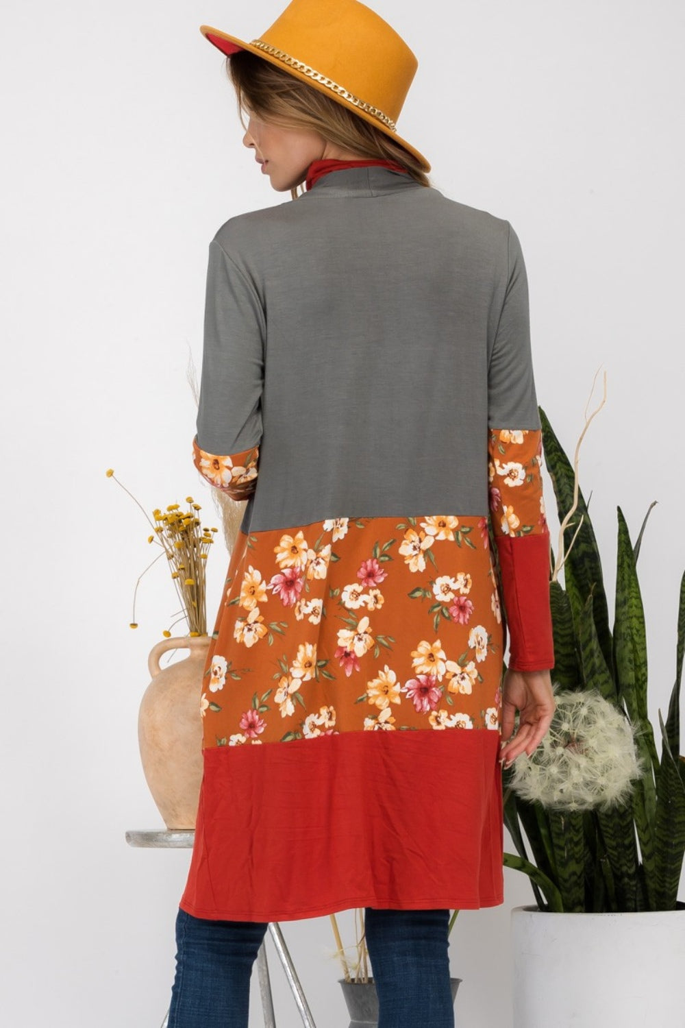 Wearing the Celeste Full Size Floral Color Block Open Front Cardigan, a person in a red hat stands indoors near potted plants, smiling.