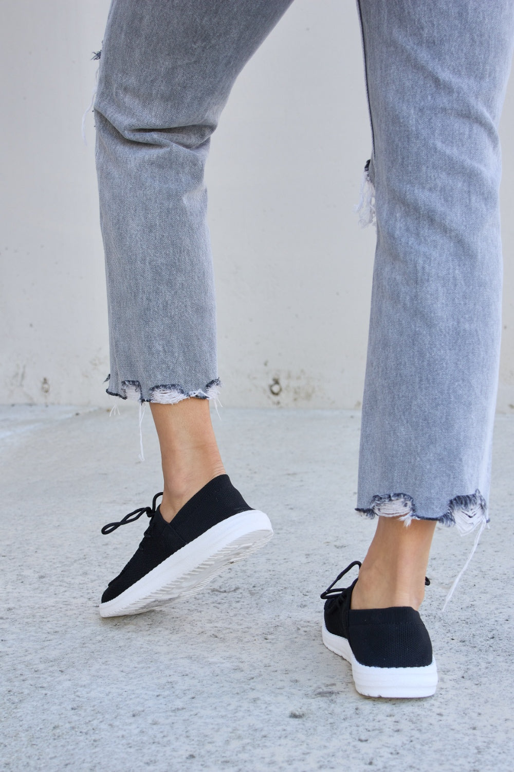 A person wearing ripped jeans and Forever Link Flat Round Toe Lace-Up Sneakers with white soles stands on a concrete surface.