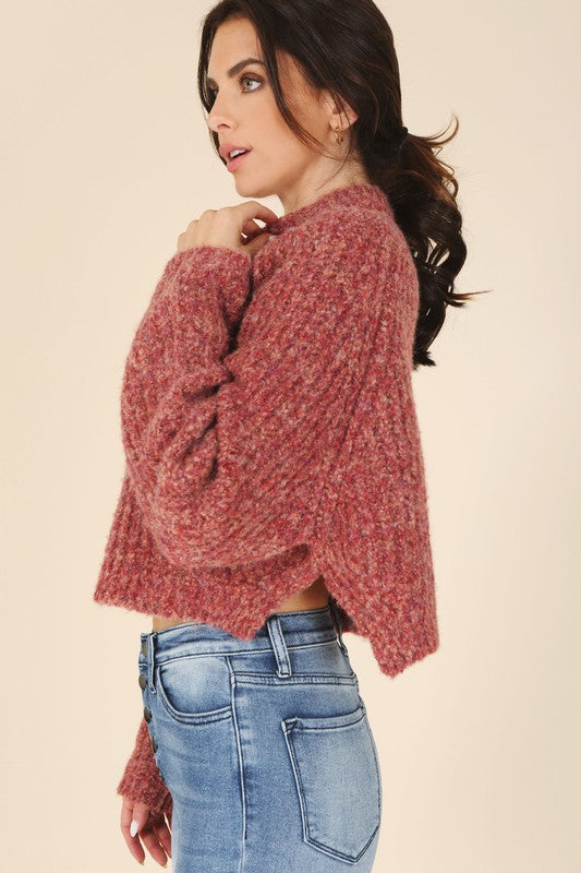 A person is wearing the Melange multicolor sweater top, characterized by textured pink fabric and featuring raglan sleeves along with large buttons, paired with light blue jeans, standing against a plain background.