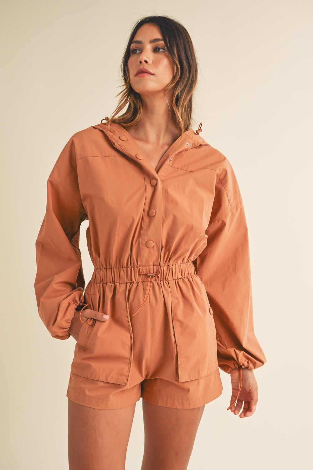 A person wearing the Mable Snap Down Long Sleeve Hooded Romper, featuring a brown color, long sleeves, a snap-down front, and an elastic waistband, is standing against a plain background.