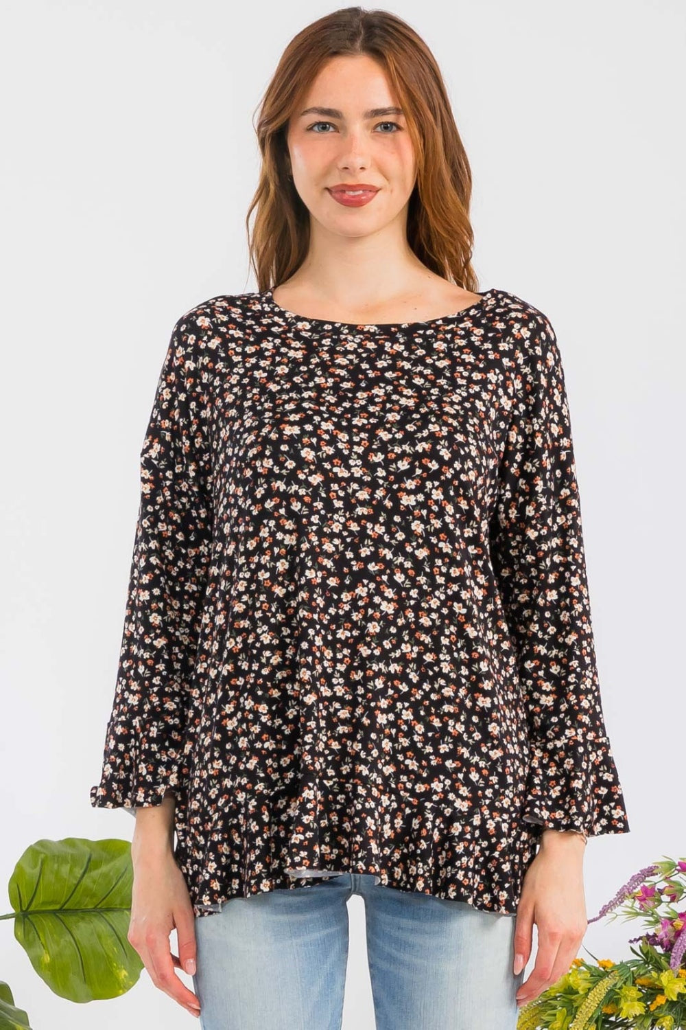 A woman elegantly stands beside colorful flowers against a plain background, embodying feminine fashion in the Celeste Full Size Floral Ruffle Detail Top, which features a round neck and floral patterns, paired with light blue jeans.