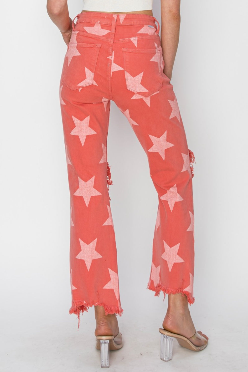 A person wearing the RISEN Full Size Distressed Raw Hem Star Pattern Jeans in a coral color, complete with distressed rips at the knees and raw frayed hems. The rock 'n' roll style is completed with clear high-heeled sandals.