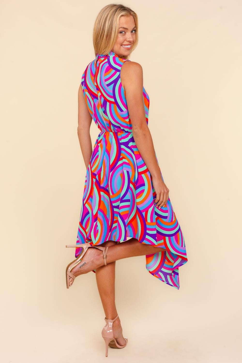 Against a beige backdrop, an individual models the Haptics Full Size Mock Neck Sleeveless Printed Dress, which features an eye-catching asymmetrical pattern. The lively blend of pink, blue, orange, and purple swirls crafts a captivating visual symphony.