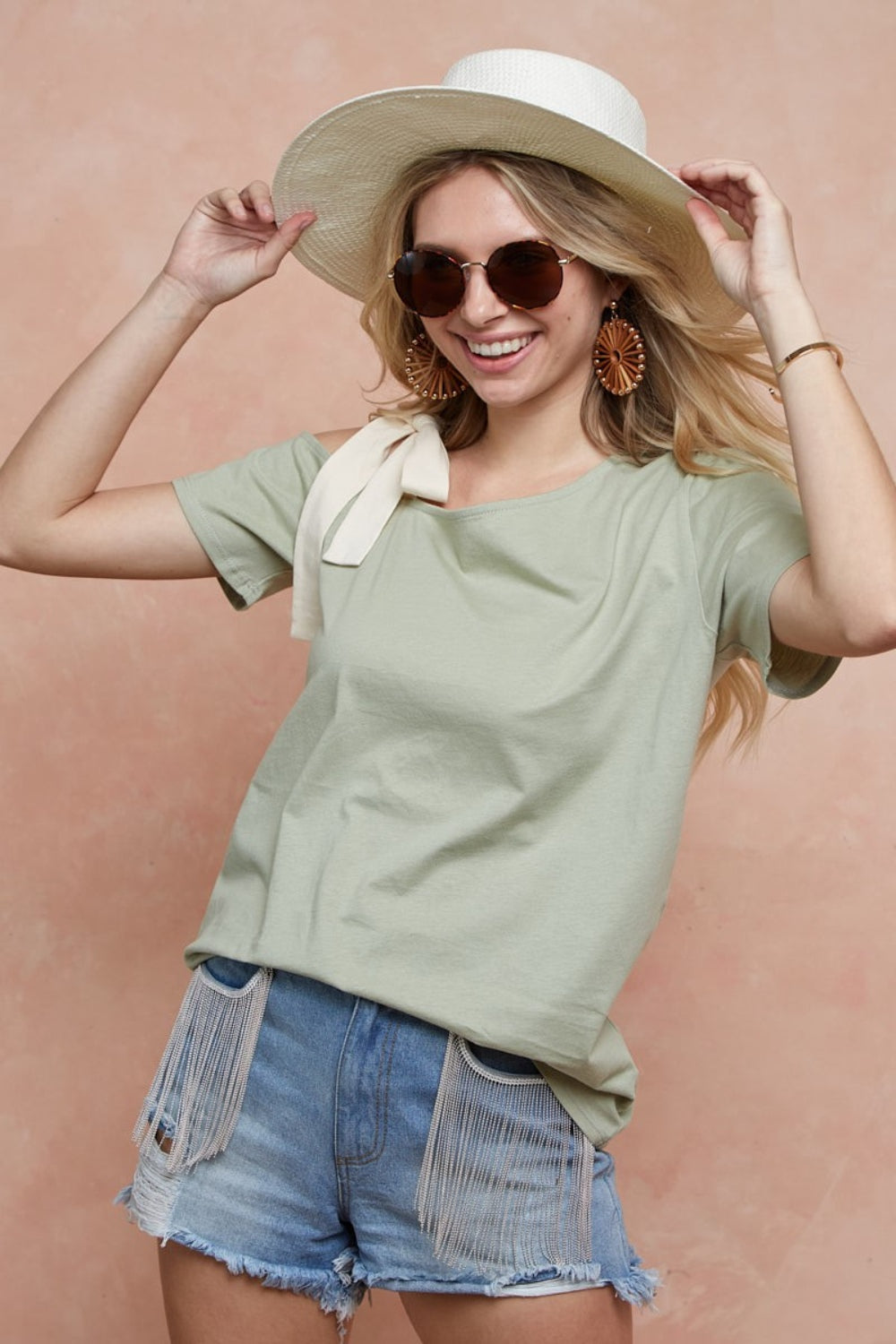 A person smiling, sporting a wide-brimmed hat, sunglasses, and the BiBi Tied Ribbon One Shoulder Short Sleeve T-Shirt in light green, paired with denim shorts, stands against a peach background.