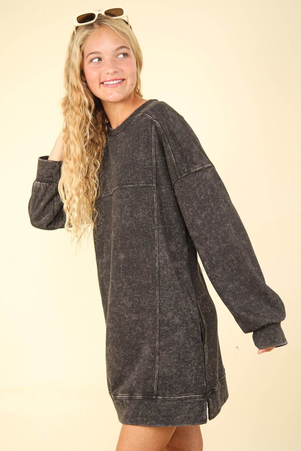 A person with long, wavy blonde hair wears a VERY J Mineral Washed Oversized Sweatshirt Mini Dress with pockets and sunglasses on top of their head, set against a beige background.