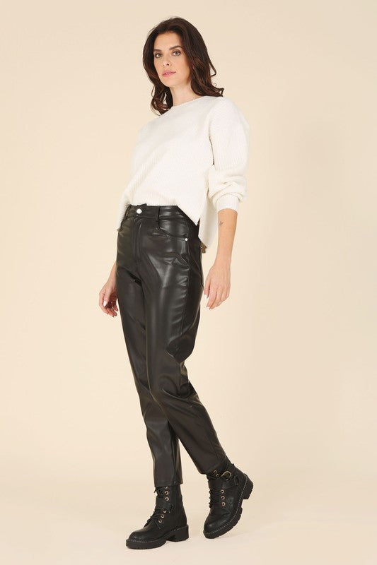 A person wearing a white sweater and black Vegan leather pants with hands in the pockets.