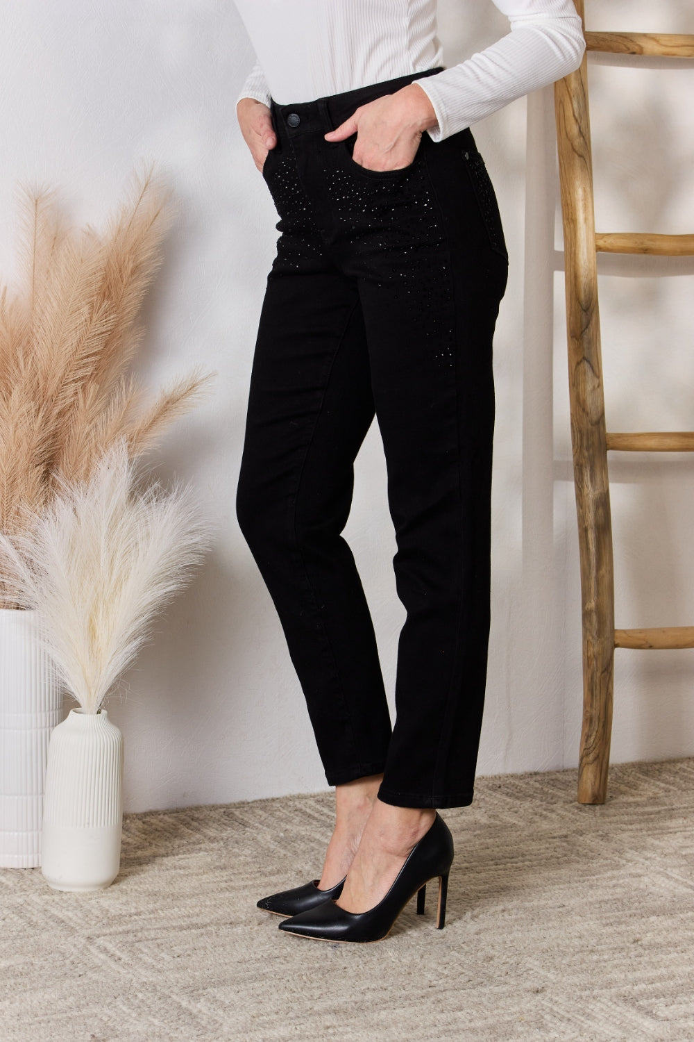 A person wearing slightly stretchy, black Judy Blue Full Size Rhinestone Embellished Slim Jeans and black shoes stands on a textured floor next to a white vase with brown and white ornamental grasses and a wooden ladder.