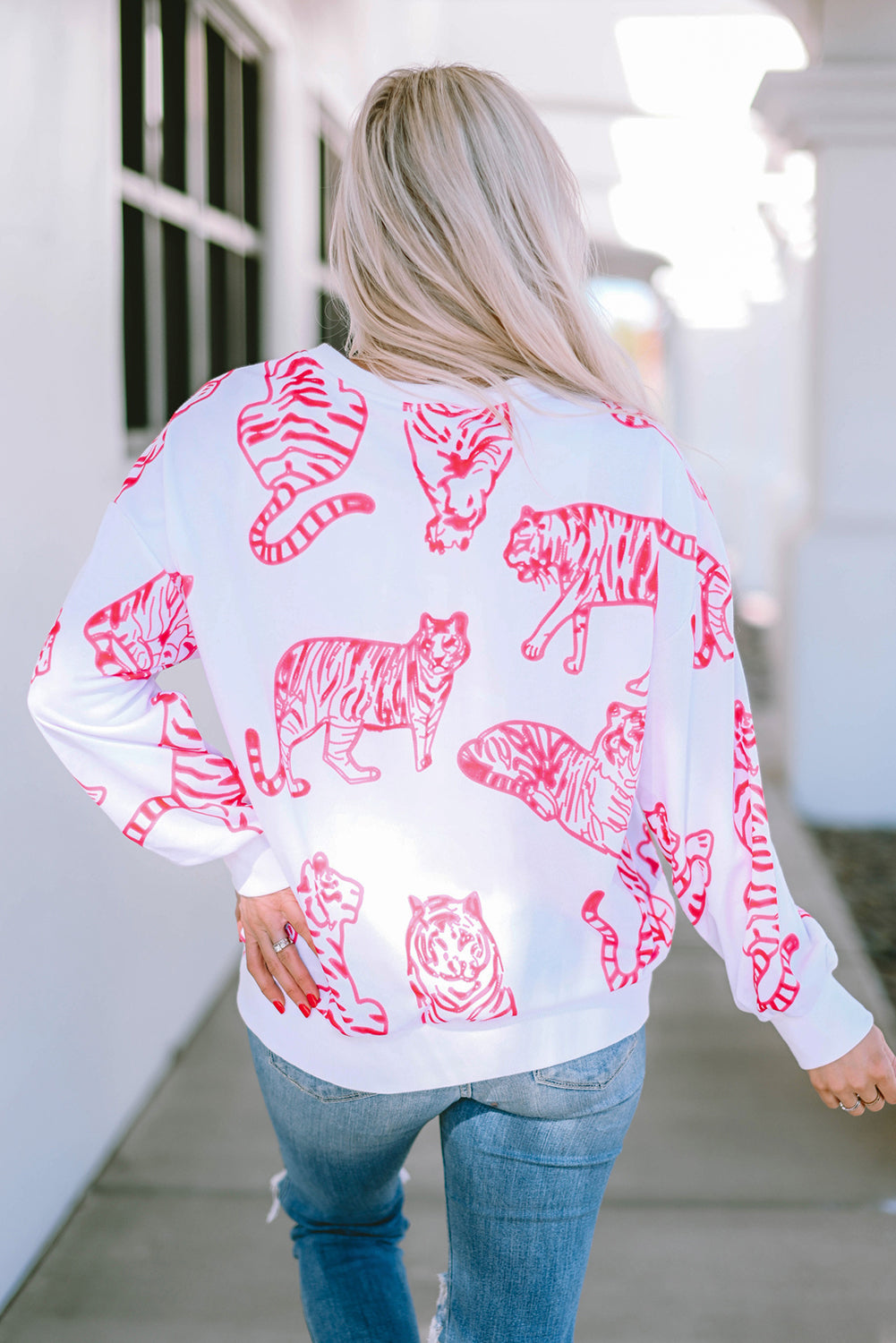 A person with long blonde hair wears a trendy Bright White Pink Animal Print Pullover Sweatshirt, paired with blue jeans, standing outdoors with their back facing the camera.