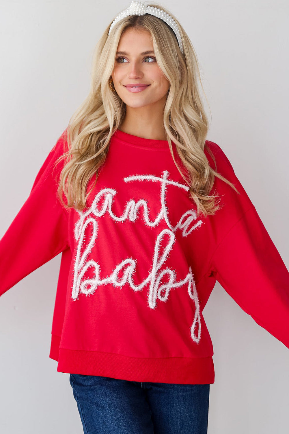A person with long blonde hair is seen wearing the Fiery Red Christmas Santa Baby Tinsel Graphic Sweatshirt and blue jeans, facing away from the camera against a plain background.