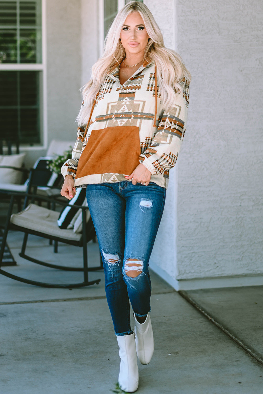 A person with long blonde hair, seen from behind, is wearing the Beige Aztec Print Kangaroo Pocket Half-Zip Hoodie. The hoodie showcases an Aztec pattern in shades of brown, beige, and orange and features a drop shoulder design with a cozy kangaroo pocket, all while standing outdoors.