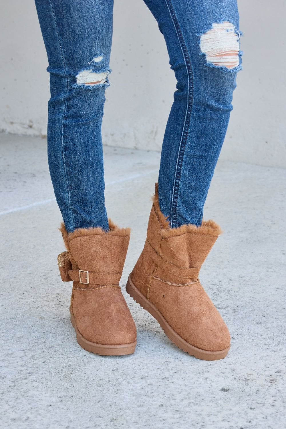 Person wearing ripped blue jeans and Forever Link Plush Thermal Flat Boots, tan in color, fur-lined with side buckles, standing on a concrete surface. These stylish boots promise cozy insulation for the feet.