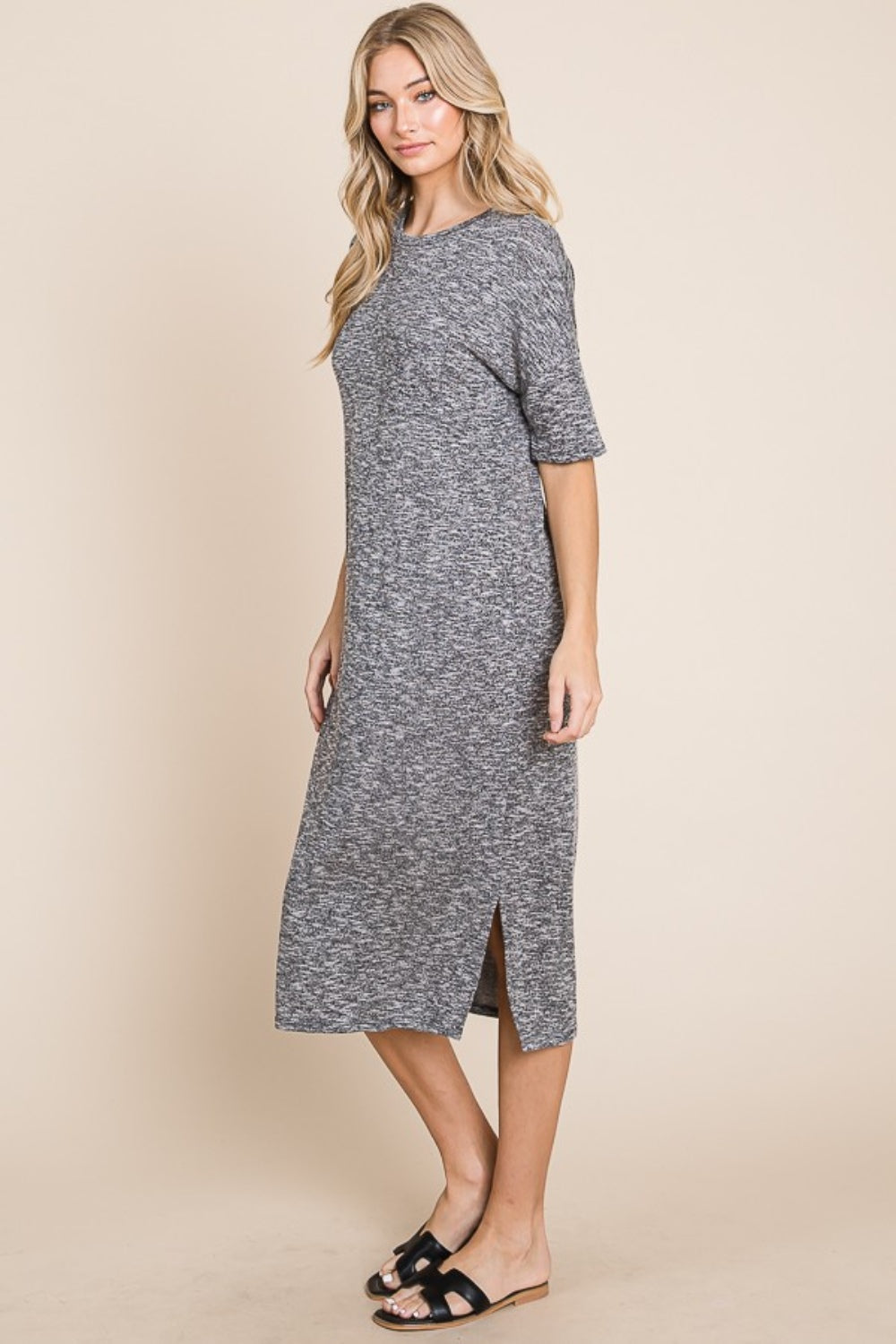 A woman wearing the BOMBOM Slit Round Neck Half Sleeve Midi Dress, a knee-length, grey midi dress featuring short sleeves and a slit detail at the hem, paired with black slip-on sandals, stands in front of a neutral background.