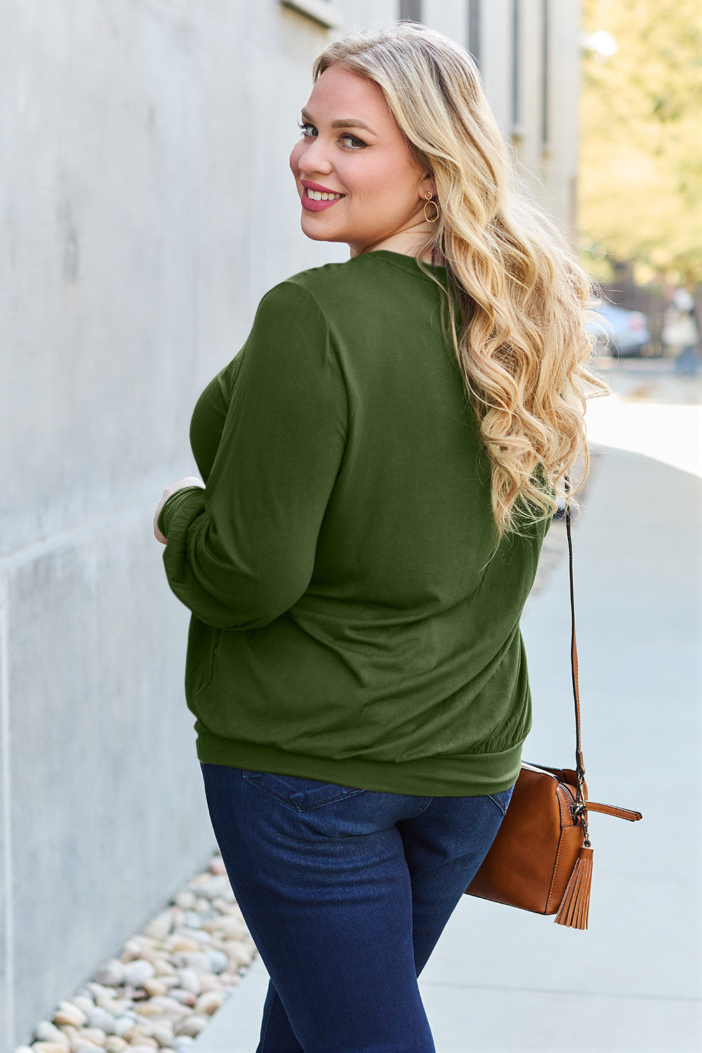 A woman with brown hair is standing against a concrete wall, wearing the Basic Bae Full Size V-Neck Lantern Sleeve Top in teal and blue basic-style jeans, smiling.