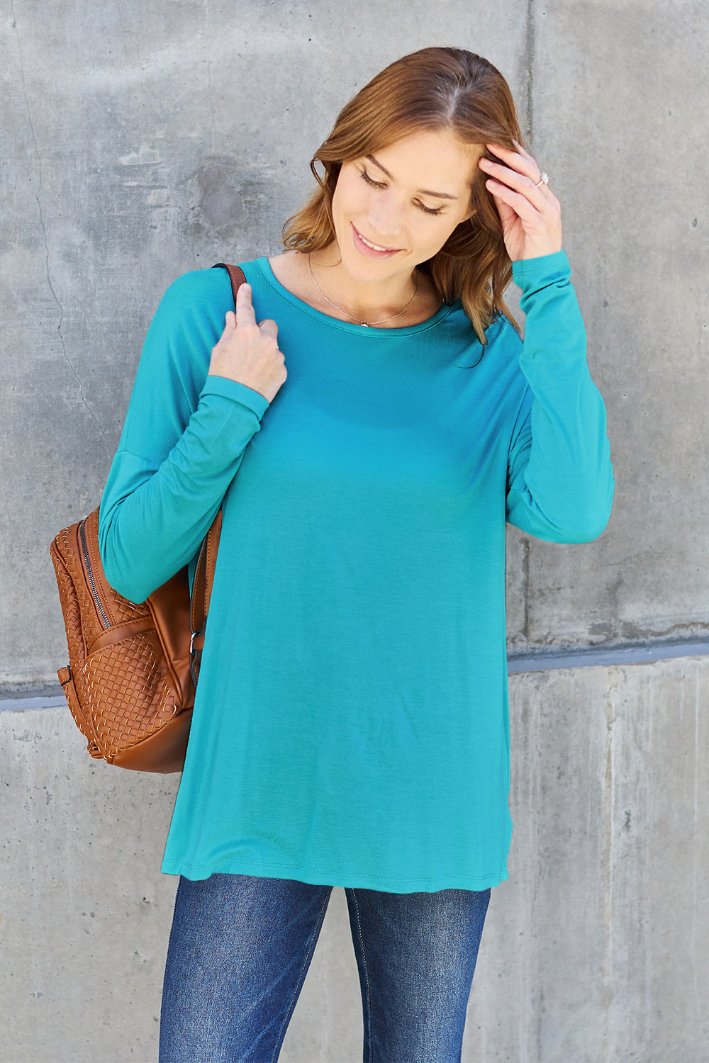 A woman with shoulder-length hair wearing a Basic Bae Full Size Round Neck Dropped Shoulder T-Shirt in green and a pair of jeans, exuding a basic style, stands against a concrete wall. She's holding an imported brown woven backpack on her shoulder while touching her hair with her hand.