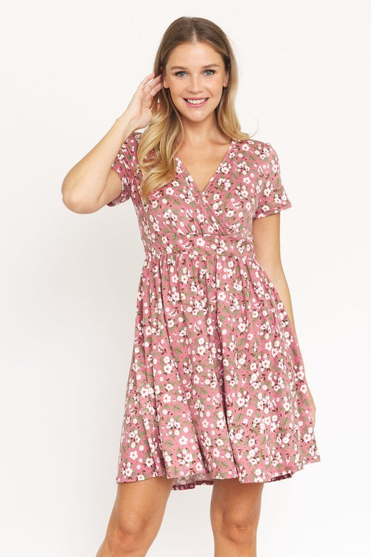 A woman wearing a short-sleeved, V-neck, Ditsy Surplice Fit and Flare Midi Dress with a dark floral background, crafted from luxurious Polyester Spandex and proudly Made in the United States, poses against a white backdrop, smiling as she softly holds the sides of her dress.