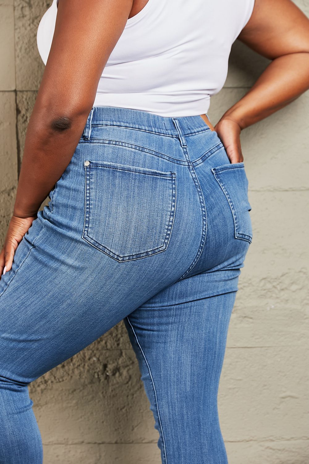 Person wearing Judy Blue Janavie Full Size High Waisted Pull On Skinny Jeans with release hem detailing and white sandals stands against a textured wall.