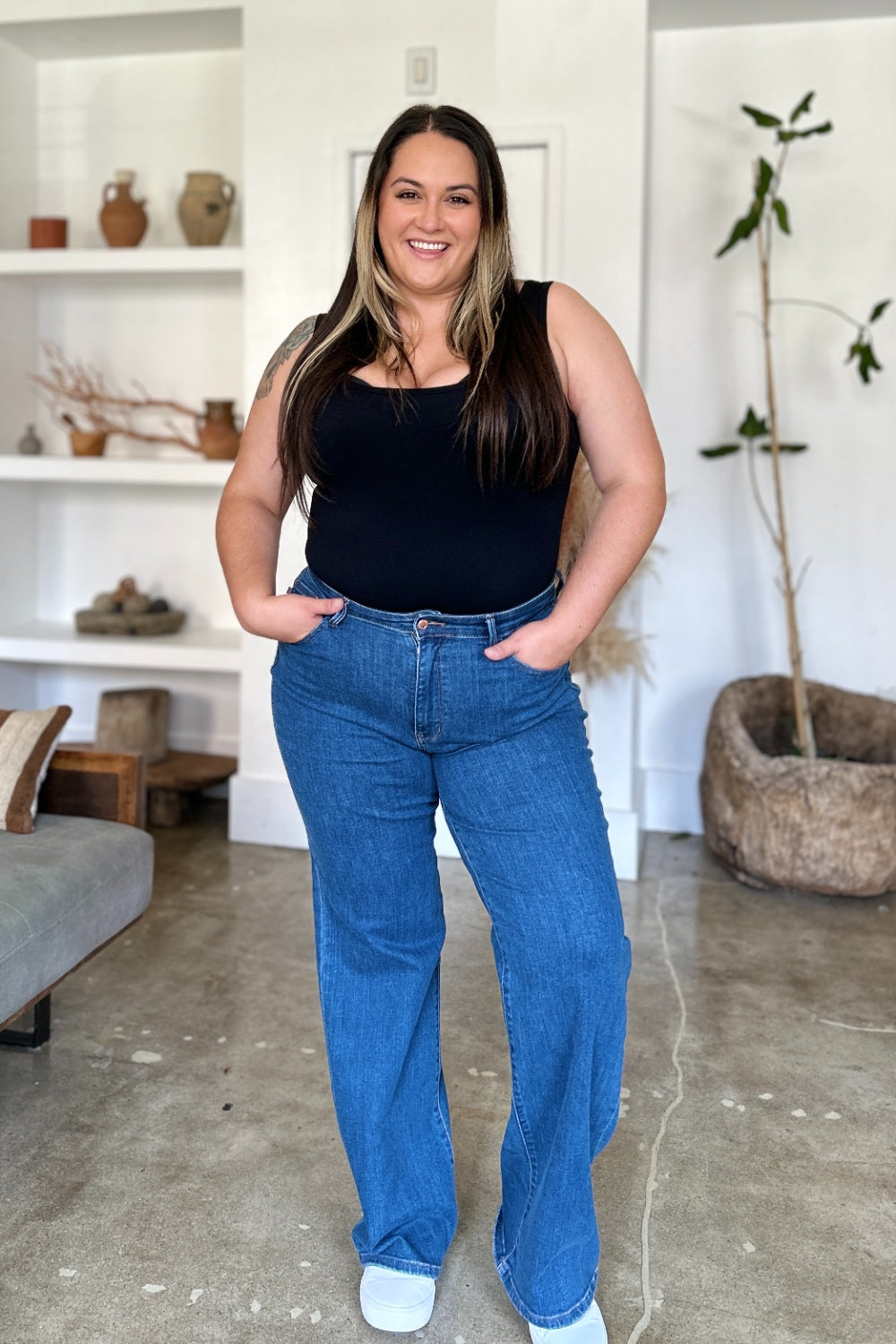 A person wearing Judy Blue Full Size High Rise Straight Jeans, a black top, and white shoes stands gracefully on a polished concrete floor.