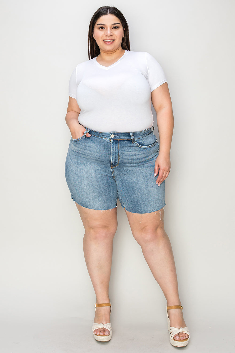 Side view of a person wearing a white shirt and Judy Blue Full Size High Waist Raw Hem Denim Shorts, standing indoors near a wooden cabinet. Summer fashion vibes radiate through the casual ensemble.