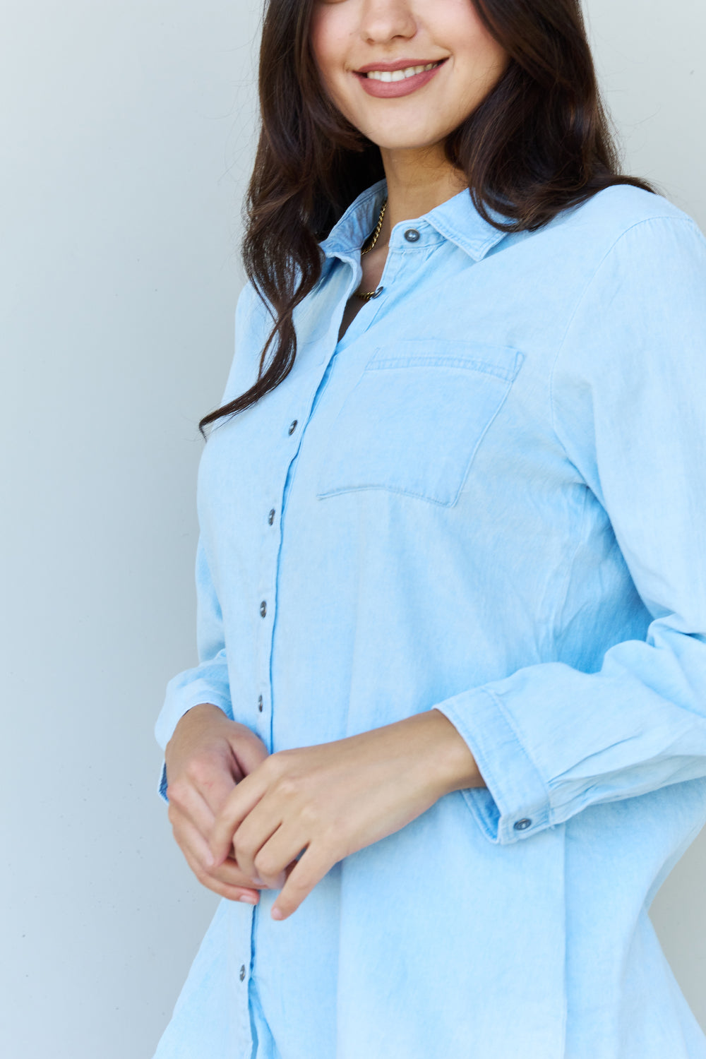 A woman wearing the Doublju Blue Jean Baby Denim Button Down Shirt Top in Light Blue, paired with white ripped shorts, strikes a pose with one hand in her pocket against a plain background, highlighting the classic button-up silhouette.