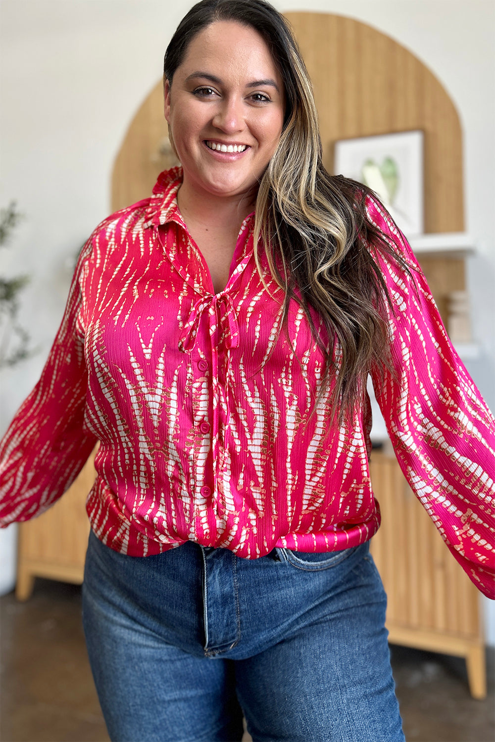 A woman wearing the Double Take Full Size Printed Button Up Long Sleeve Shirt in vibrant pink polyester and black pants stands against a plain background while touching her hair.