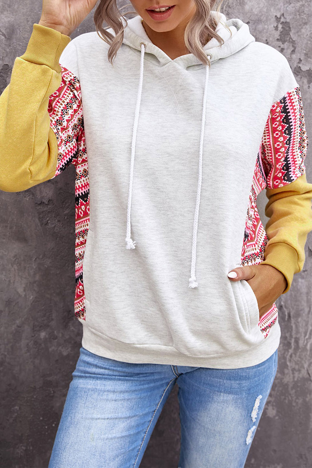 A person with long hair wearing an Aztec Patch Pullover Hoodie and blue jeans is facing away from the camera against a textured gray wall.