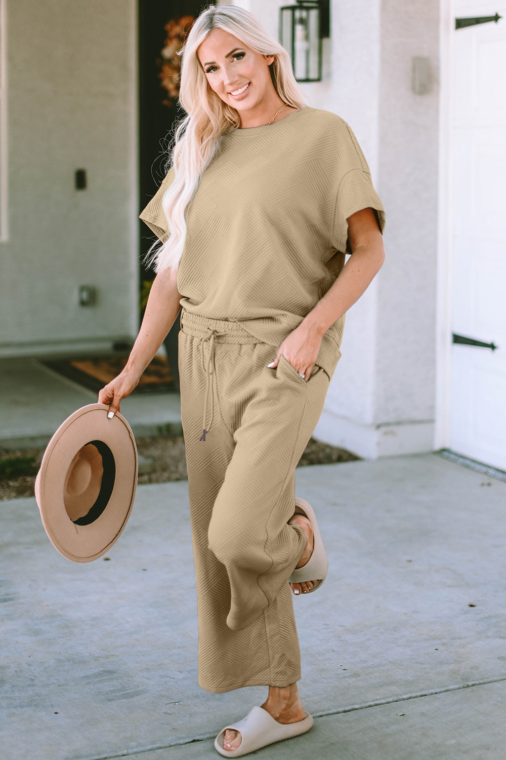 A woman with long hair smiles while standing outside a building, dressed in the Double Take Full Size Texture Short Sleeve Top and Pants Set, featuring a matching beige two-piece ensemble. She wears slides and holds a hat in her right hand. Her outfit, slightly stretchy for added comfort, is perfect for machine wash cold.