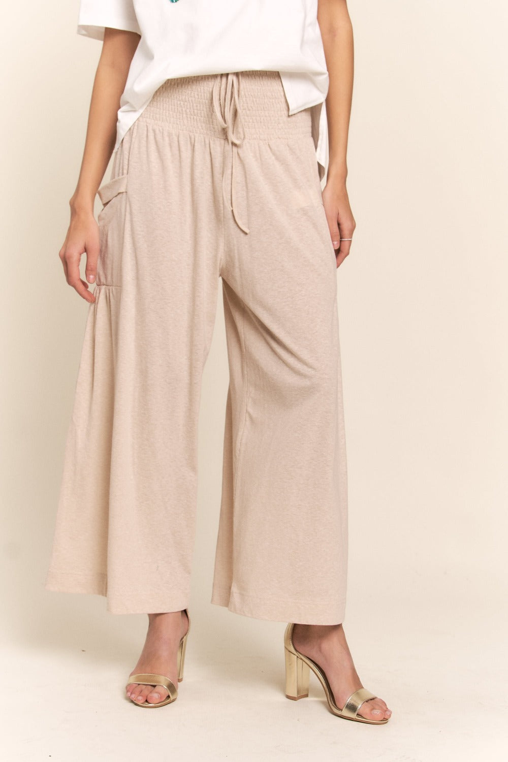 A person wearing J.NNA Smocked Waist Boho Wide Leg Pants with Pockets in beige stands in low-heeled sandals against a plain background, paired with a chic white T-shirt.