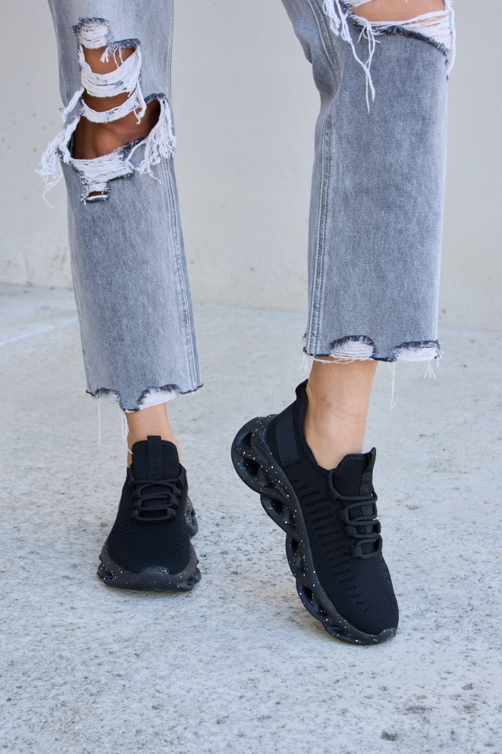 A person wearing ripped gray jeans and Forever Link Mesh Breathable Lace-Up Athletic Shoes is standing on a concrete surface.