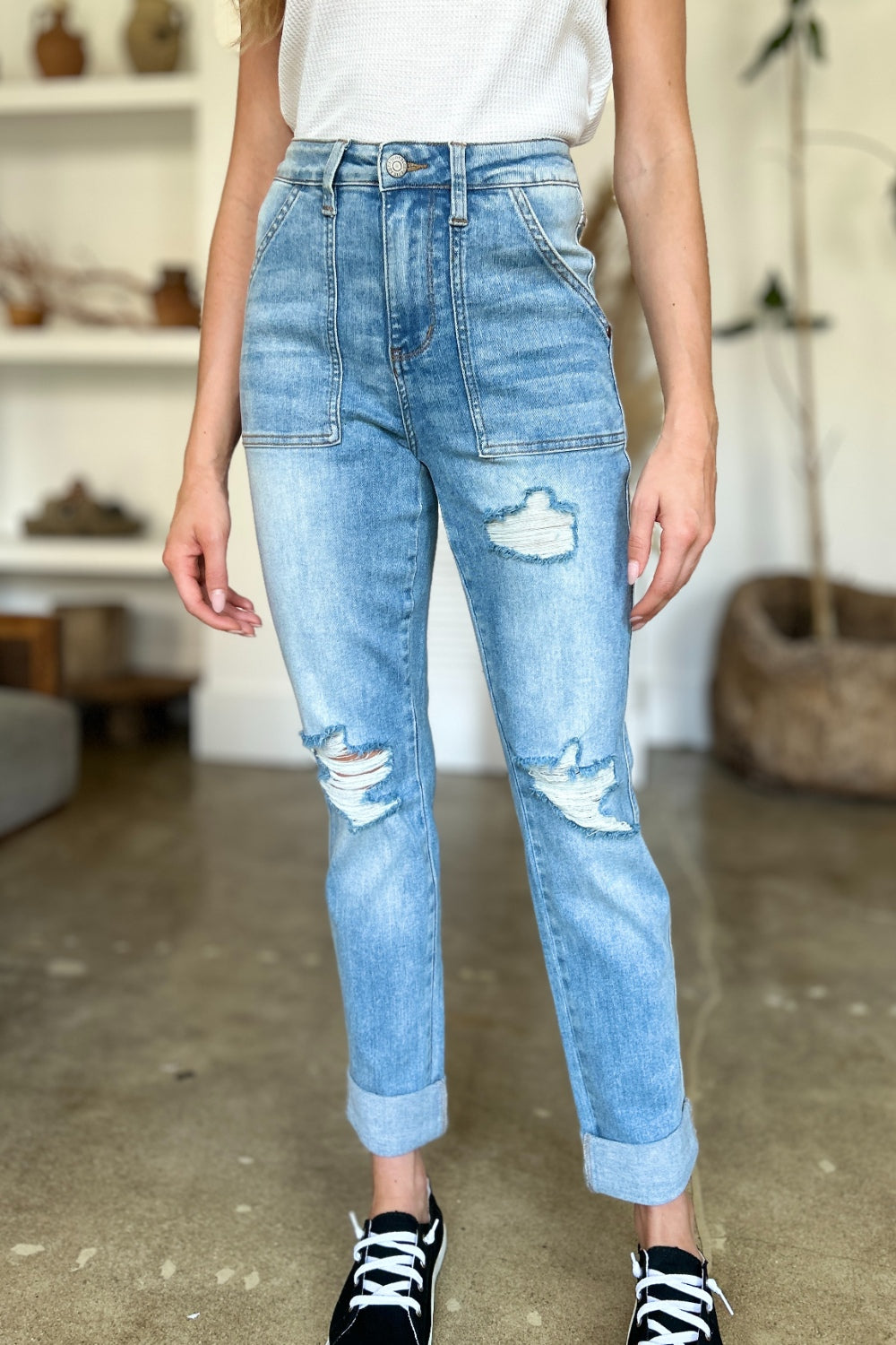 Two women are standing side by side, both clad in black and white tank tops and Judy Blue Full Size Distressed Straight Jeans with Patch Pockets, smiling at the camera in a modern living room.